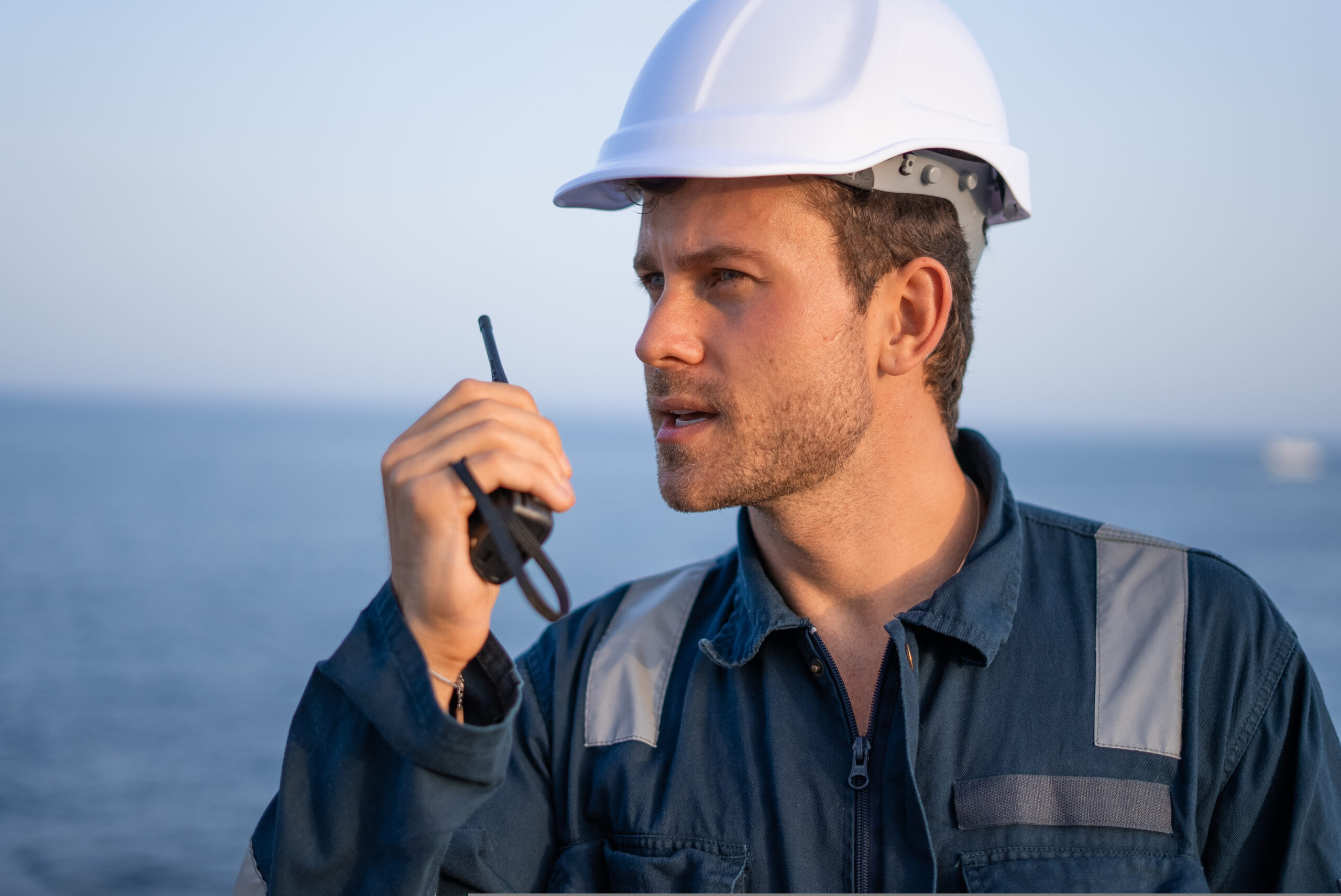 Serious young bearded male specialist in uniform and protective hardhat talking to walkie talkie while working at sea port